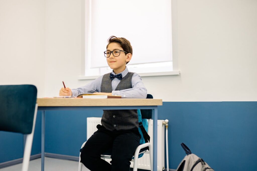 pexels-photo-8617989 A confident boy wearing glasses and a bow tie sitting at a school desk, ready to learn.